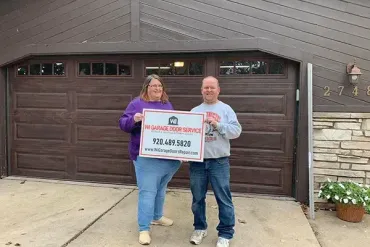 Two people stand in a driveway holding a sign for MR GARAGE DOOR SERVICE in front of a dark brown garage door.