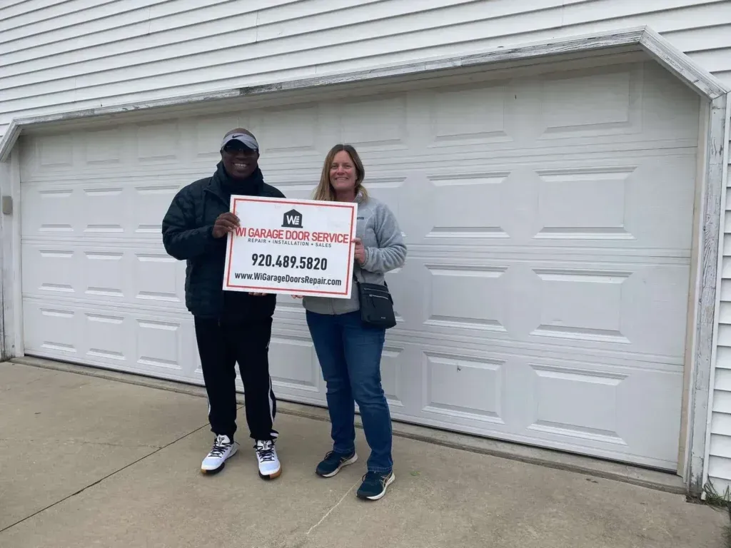 Two people stand before a white garage door, smiling while holding a sign for The Garage Door Company with phone number.