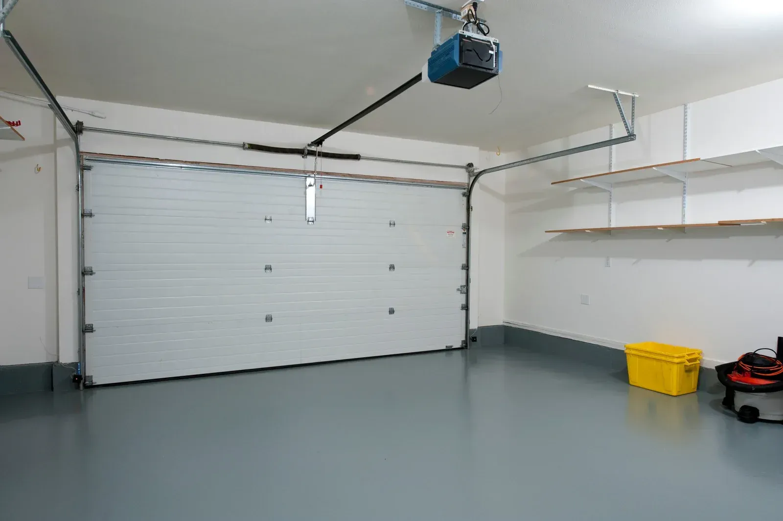 Empty white garage with closed door, gray floor, wall shelves, and a yellow bin on the right.