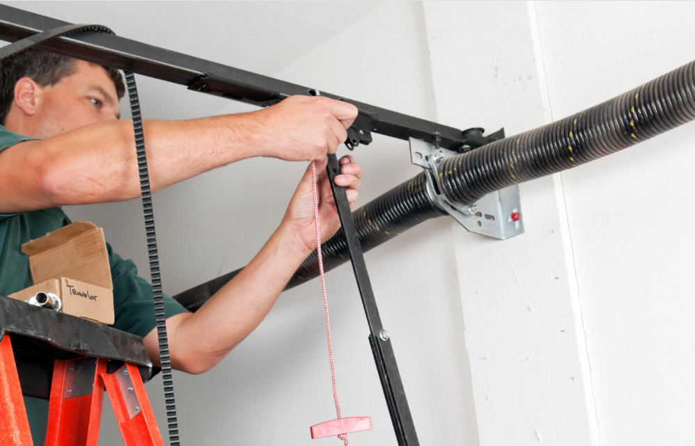 Worker installing a garage door opener on a ceiling track, using a drill beside an orange ladder.