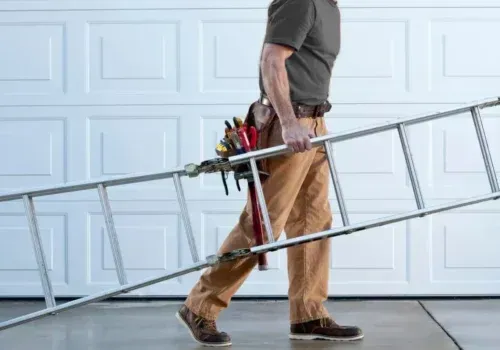 A person in work clothes and a tool belt carries a metal ladder horizontally in front of a white garage door.