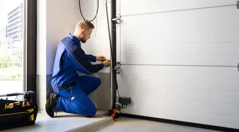 A technician in a blue uniform kneels on the floor, repairing the track of a white sectional garage door.
