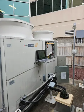 A technician’s laptop connected to a Mitsubishi outdoor HVAC unit for maintenance or diagnostic testing.