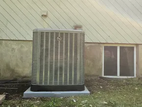 A rectangular outdoor air conditioning unit sits on a concrete pad against a house exterior wall next to a window.