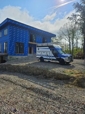 A blue, two-story house under construction with a white utility van parked in front on a gravel lot.