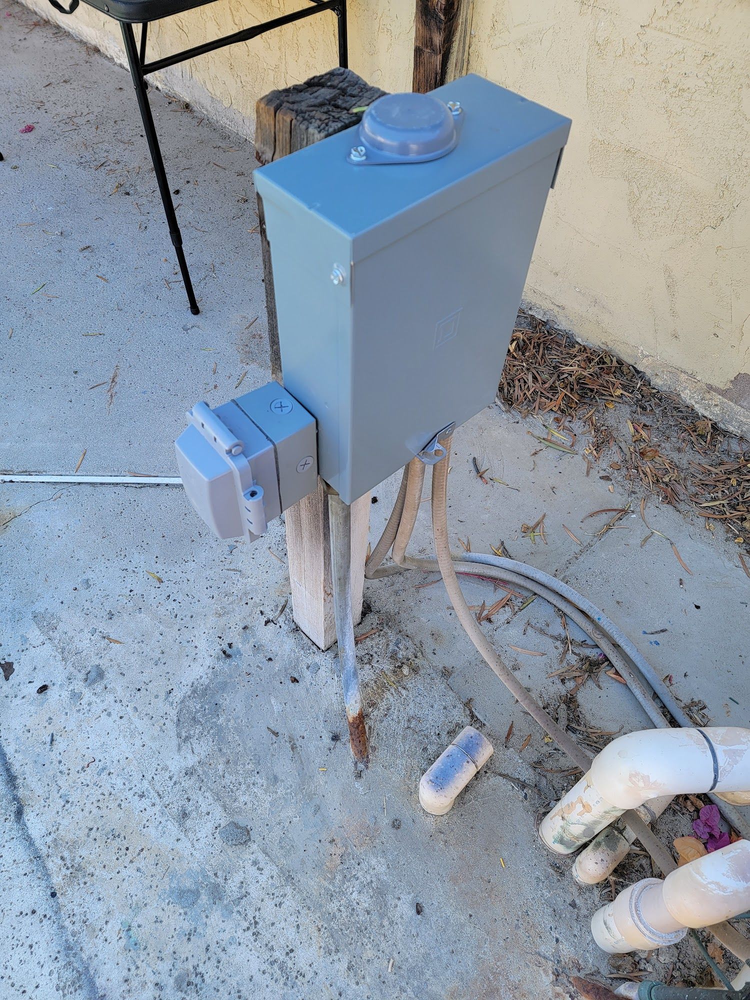Gray electrical box mounted on a wooden post beside concrete, with cables and PVC pipes nearby