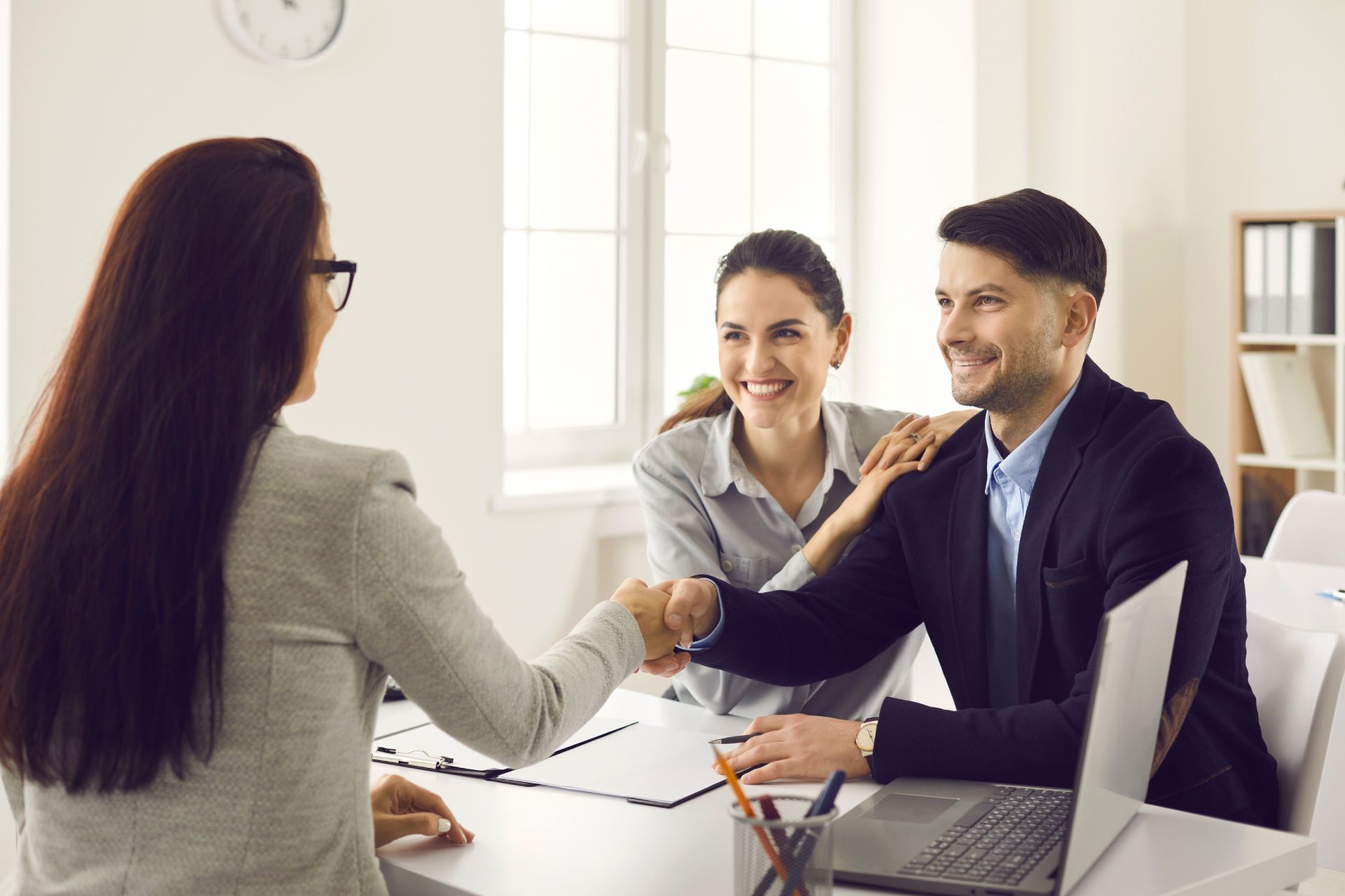 A woman shakes hands with a smiling man, with a woman in the background. They are in an office.