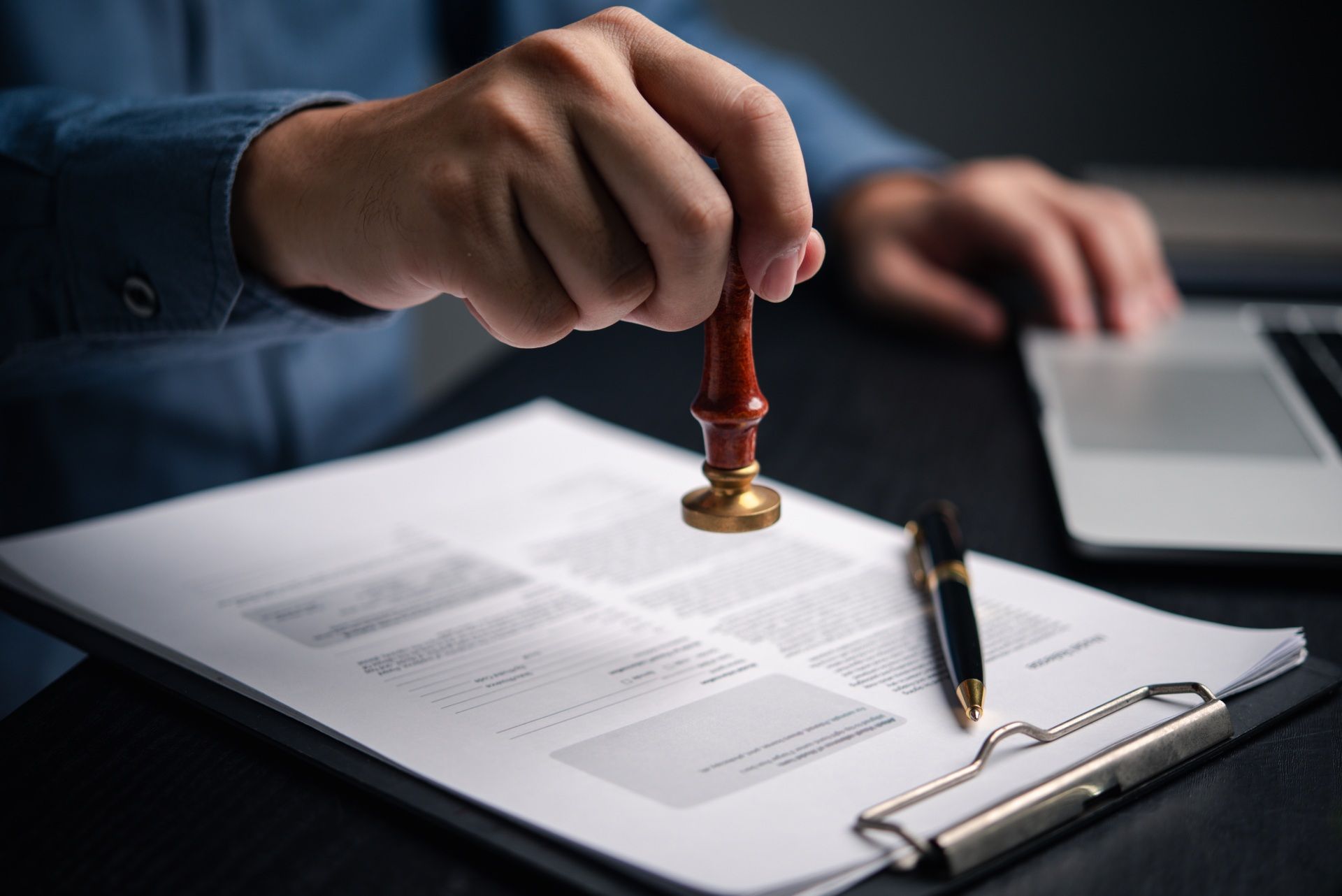 Person stamping a document with a wax seal; a laptop and pen are on the desk.