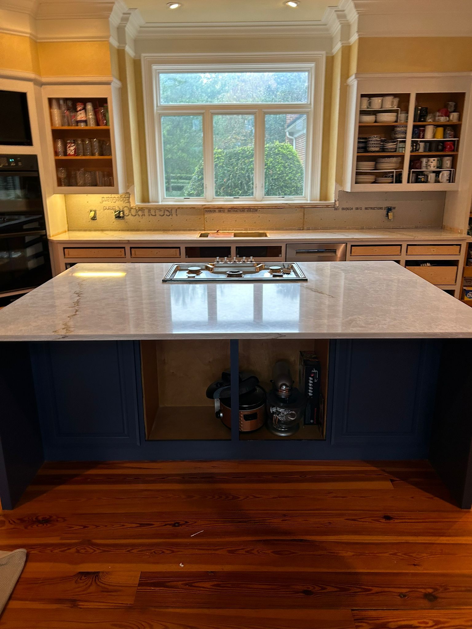 Kitchen with blue island, light countertops, and cabinets. Window centered with yellow drapes. Wooden floor.
