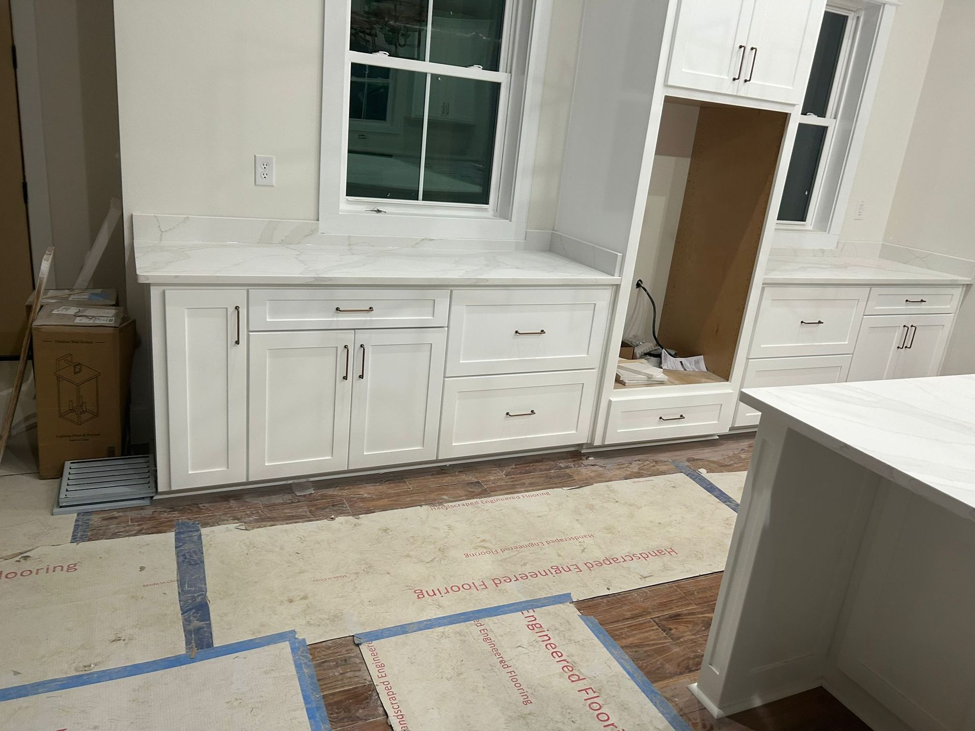 White kitchen cabinets under a window, with a countertop and island in a construction setting.