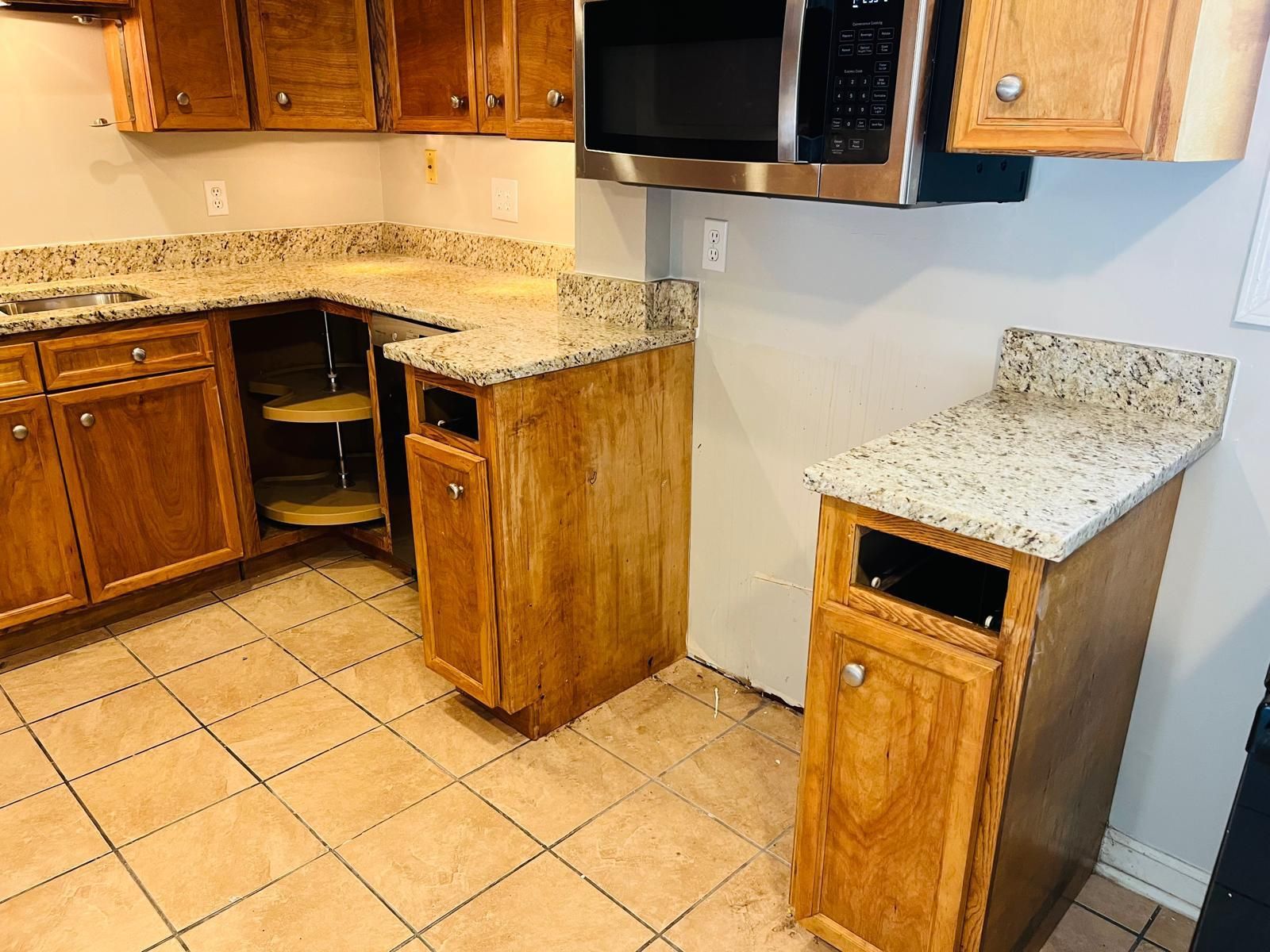 Kitchen with tan tile flooring, wooden cabinets and granite countertops. A microwave is over a counter.
