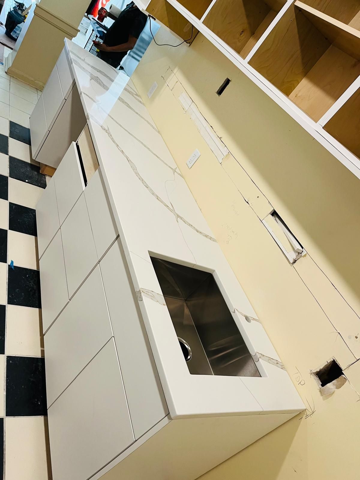 White kitchen cabinets with a sink installed, being built. Black and white checkered floor.