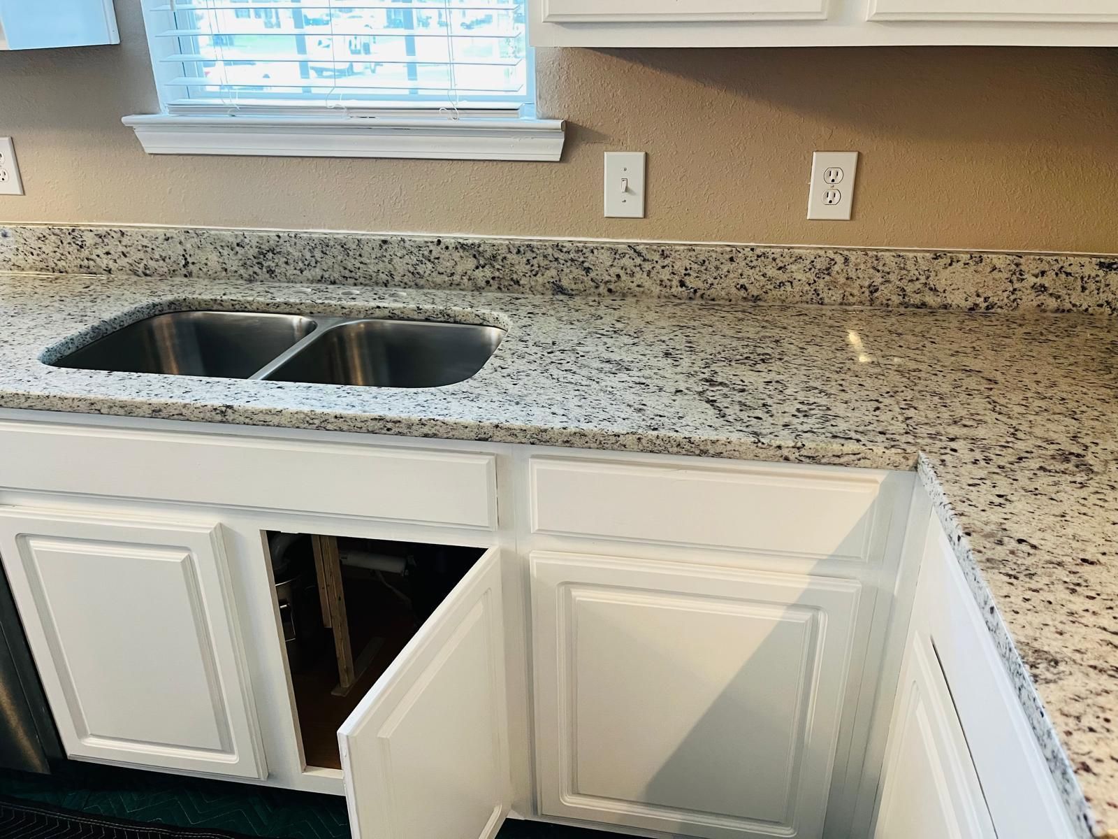 White kitchen cabinets with a granite countertop and a stainless steel sink. One cabinet door is open.