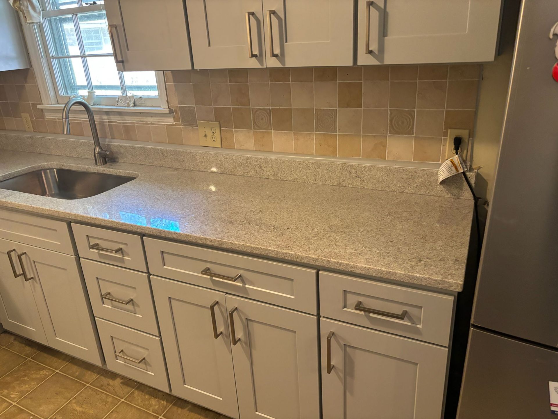 Gray kitchen cabinets with a light gray countertop, a stainless steel sink, and a light brown backsplash.
