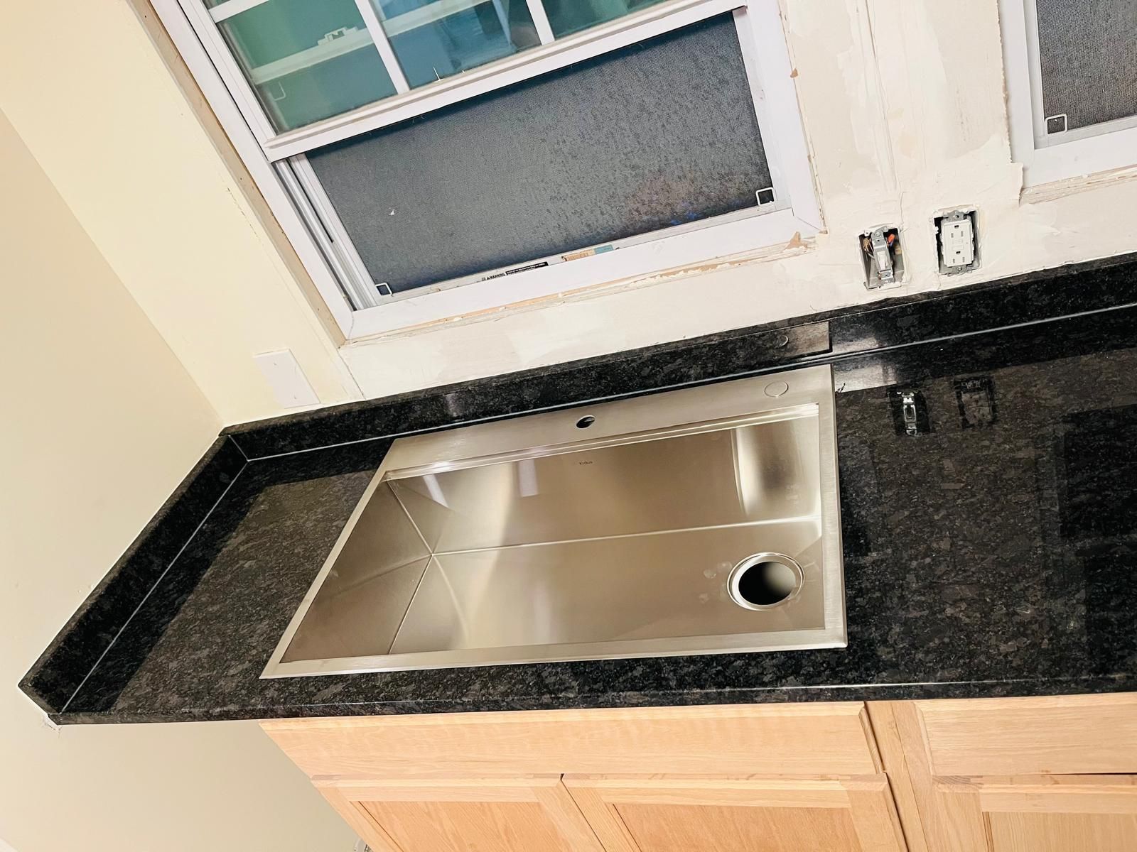 Stainless steel sink in a black granite countertop over wooden cabinets, near a window.
