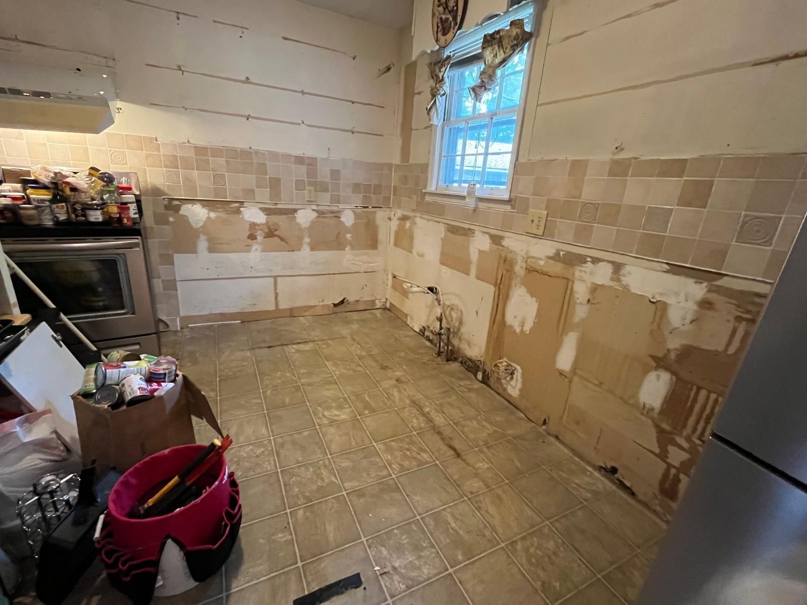 Kitchen undergoing renovation; tile removed from walls, exposing drywall. Brown floor tiles.
