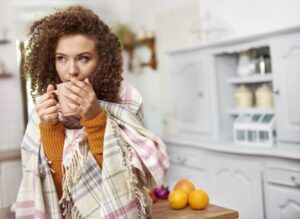 A woman wrapped in a blanket is drinking a cup of tea in a kitchen.