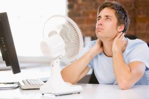 A man is sitting at a desk with a fan and a computer.