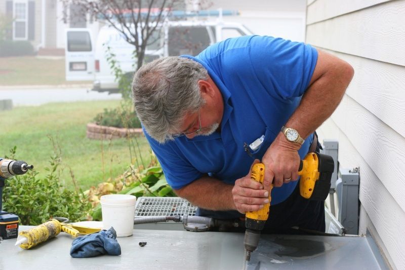 A man in a blue shirt is working on an air conditioner with a drill.