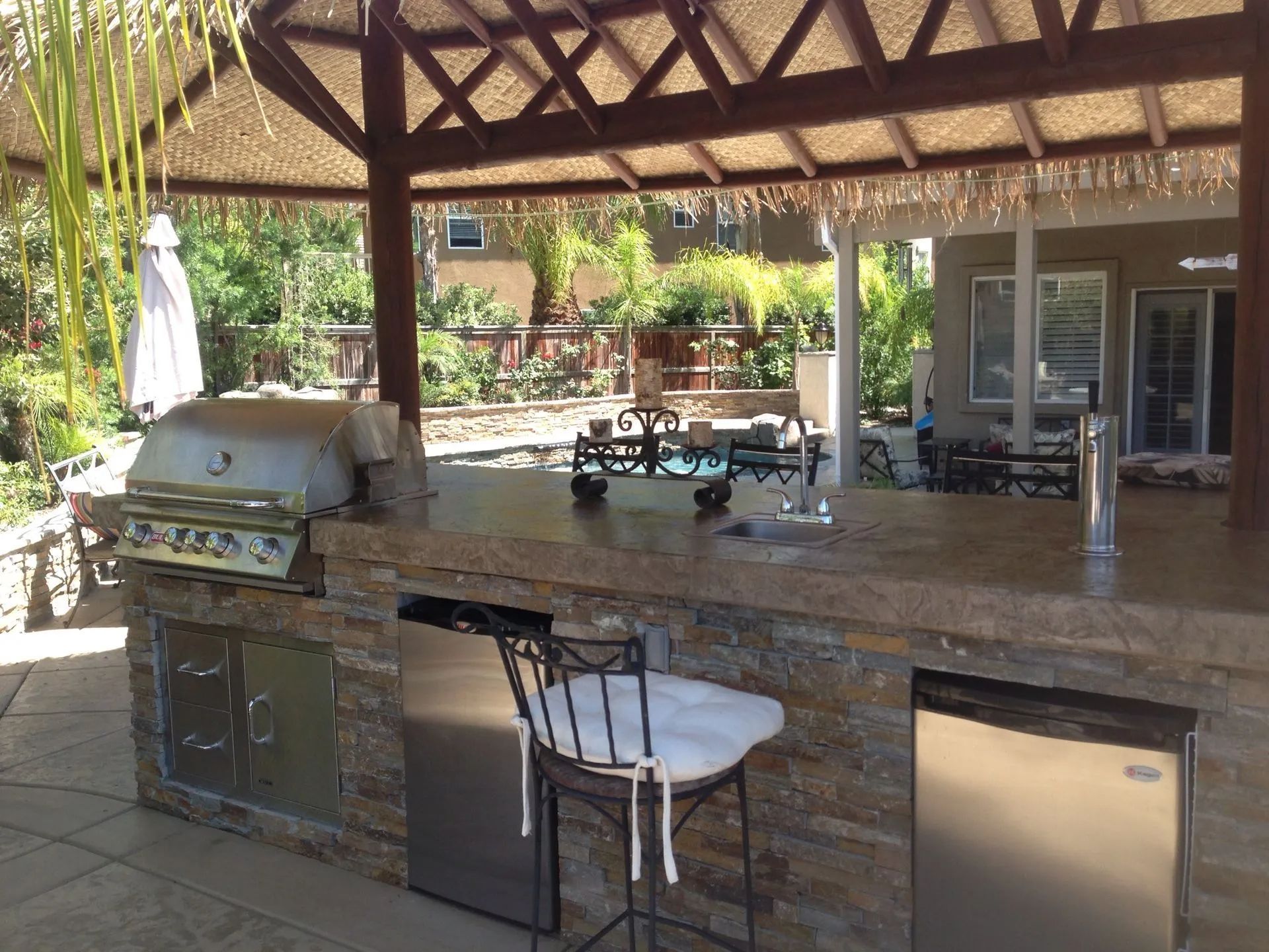 Outdoor kitchen with stone counter, grill, fridge, and tiki-style roof. A bar stool sits in front.
