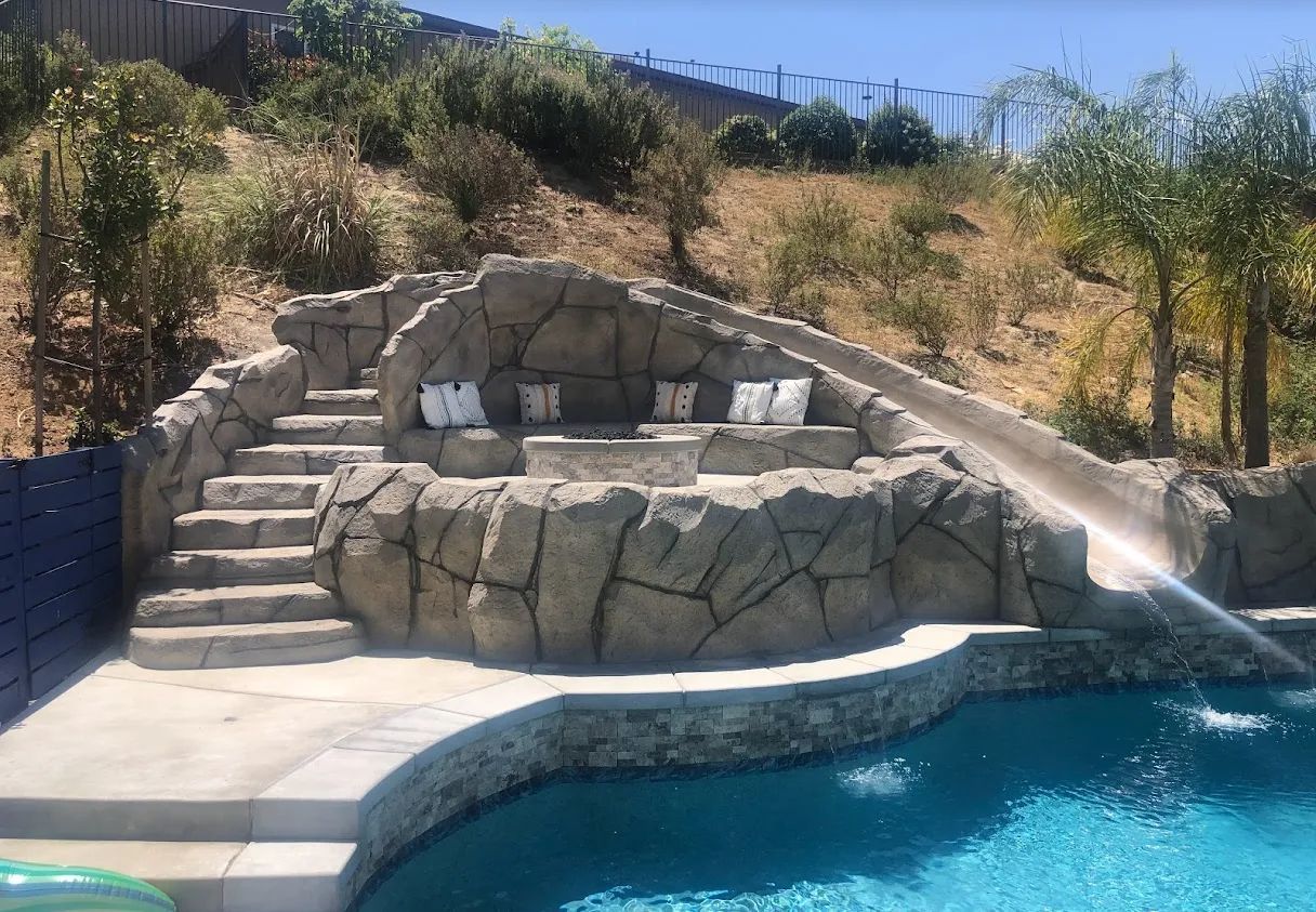 Pool with rock waterfall feature, slide, stairs, and seating area; blue water.