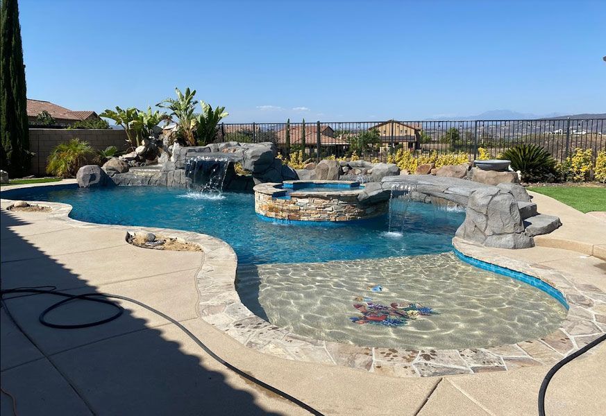 Pool with waterfall, spa, and rock features under a blue sky, surrounded by concrete patio and landscaping.