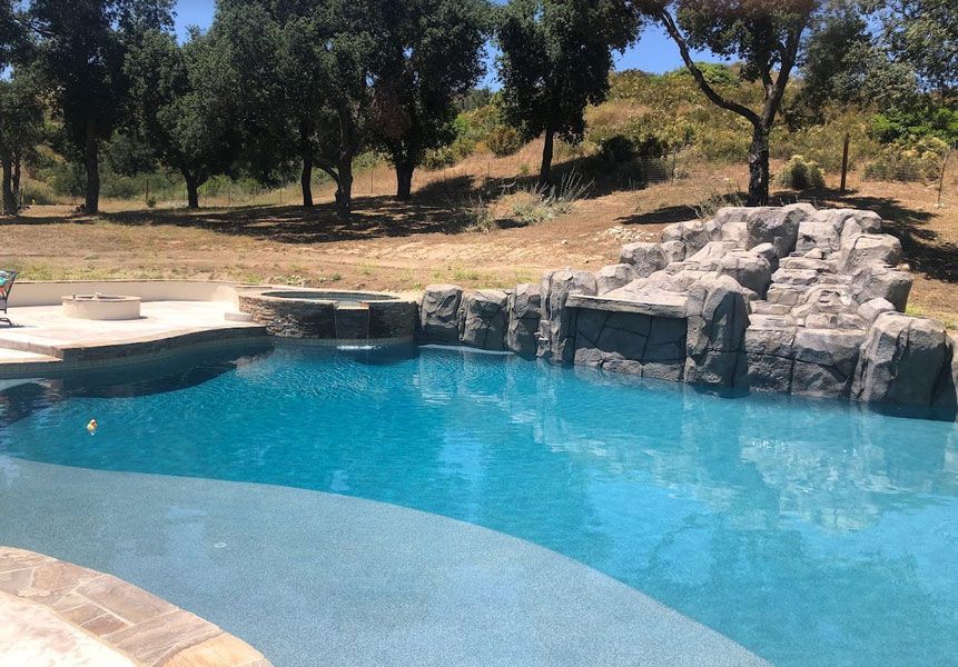 Swimming pool with waterfall feature and spa, set in a sunny, outdoor landscape. Blue water, rock detail.