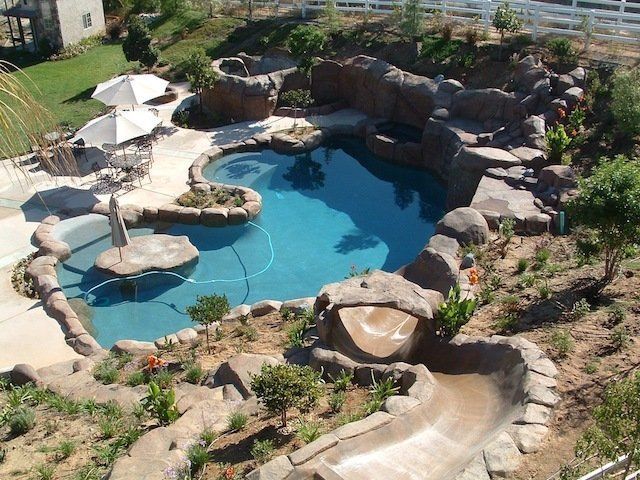 Backyard pool with rock features, slide, and patio. Blue water, brown rock, green plants, and white umbrellas.