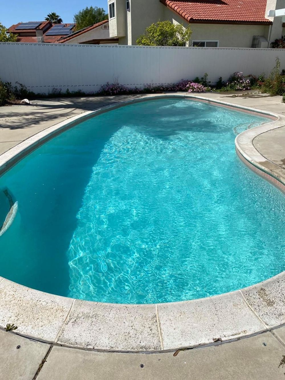 A turquoise swimming pool with a curved edge, surrounded by concrete. White wall and houses in background.