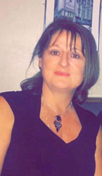 Woman with bangs, wearing a black top and pendant necklace, in front of a blurry background.