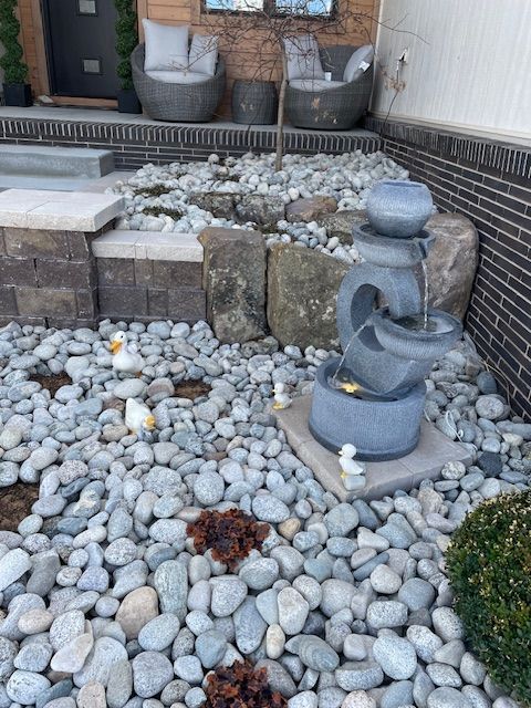 Stone fountain in a rock garden with decorative ducks and chairs in the background.