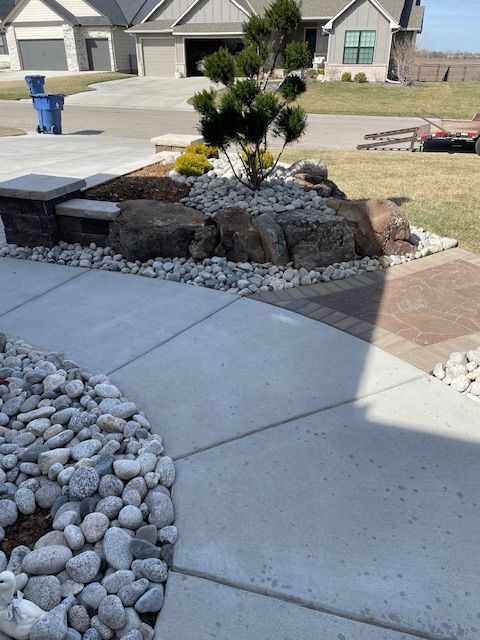 Concrete walkway leading to a landscaped rock garden with a small tree; houses in the background.