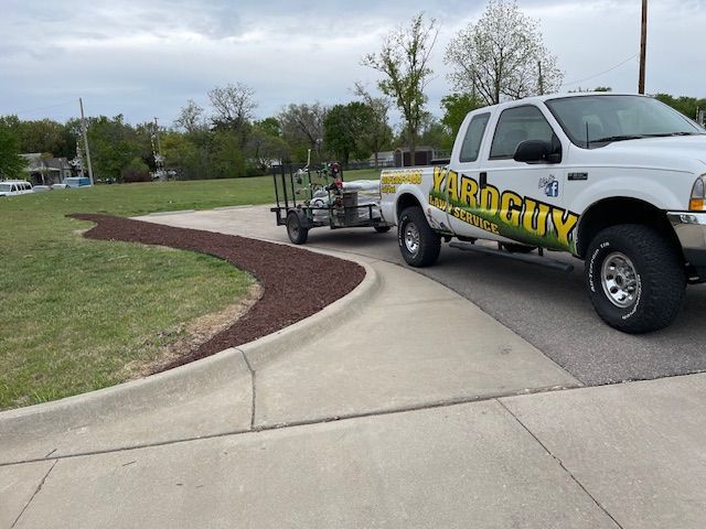 White pickup truck with trailer on a concrete driveway near a landscaped area with dark brown mulch.