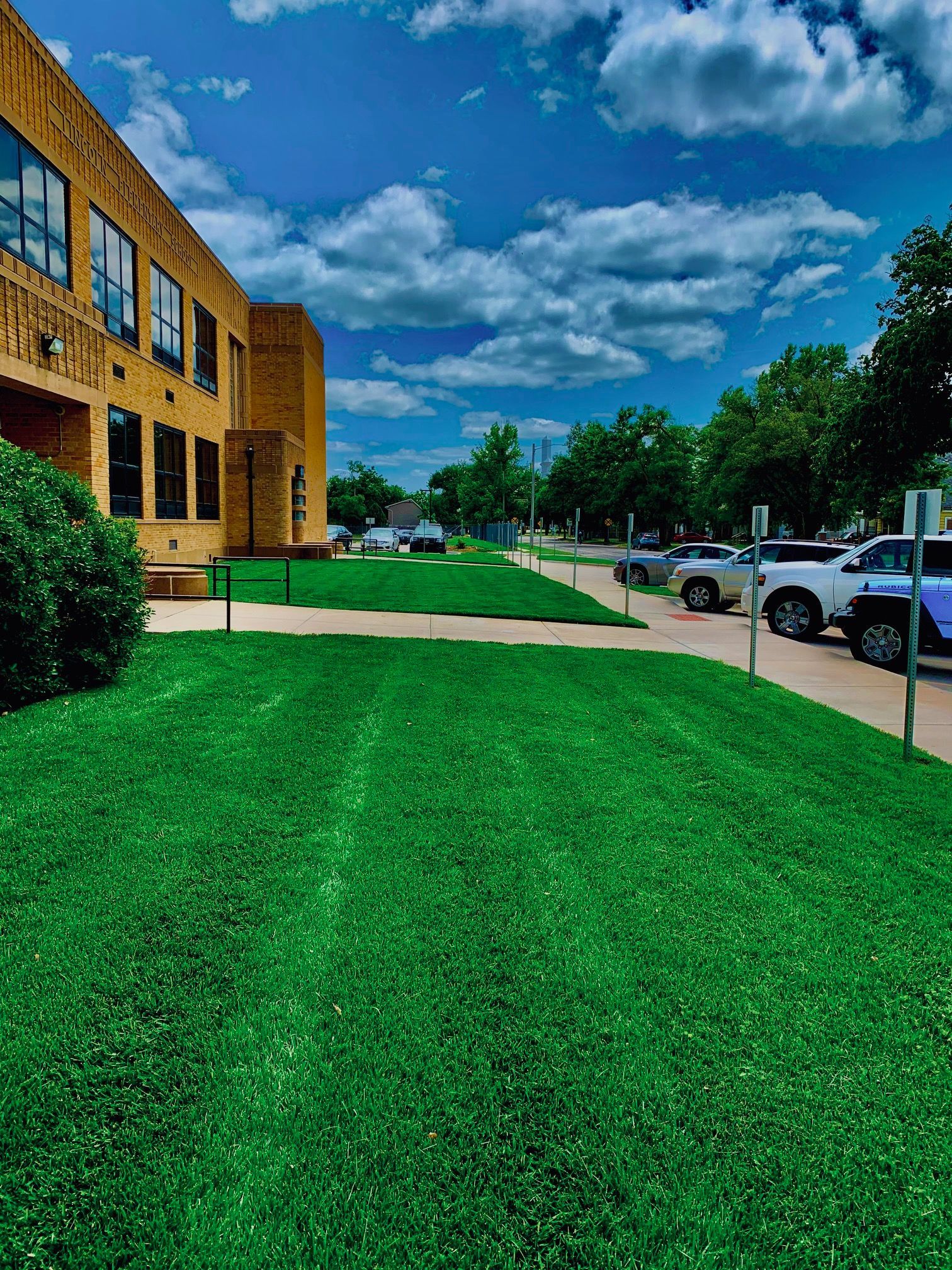 Bright green lawn in front of a tan brick building, with a cloudy blue sky overhead. Cars parked on the street.
