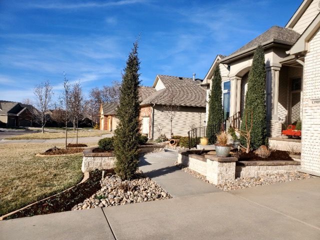 Stone-fronted house with a paved pathway, landscaping, and evergreen trees on a sunny day.