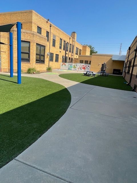 Schoolyard with artificial turf, concrete path, tan brick building, and blue poles.