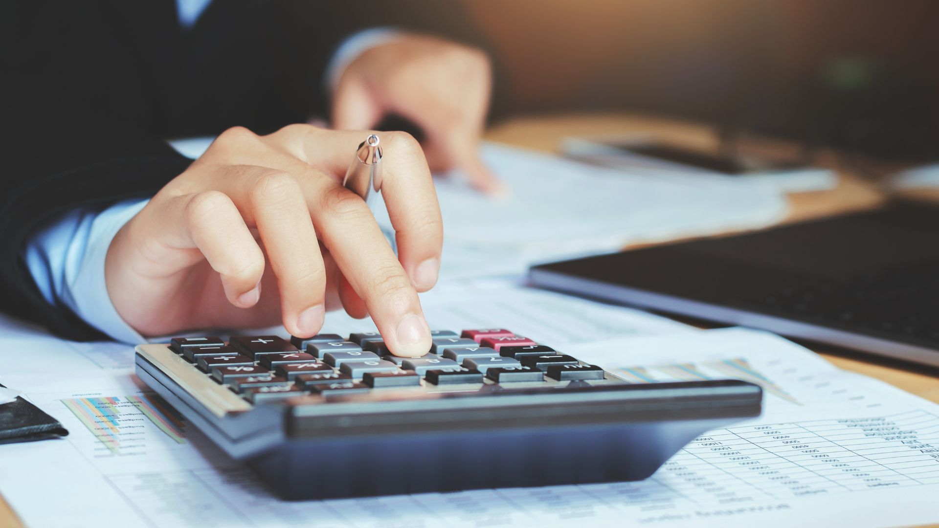 Person's hand using calculator on a desk with paperwork and a laptop.