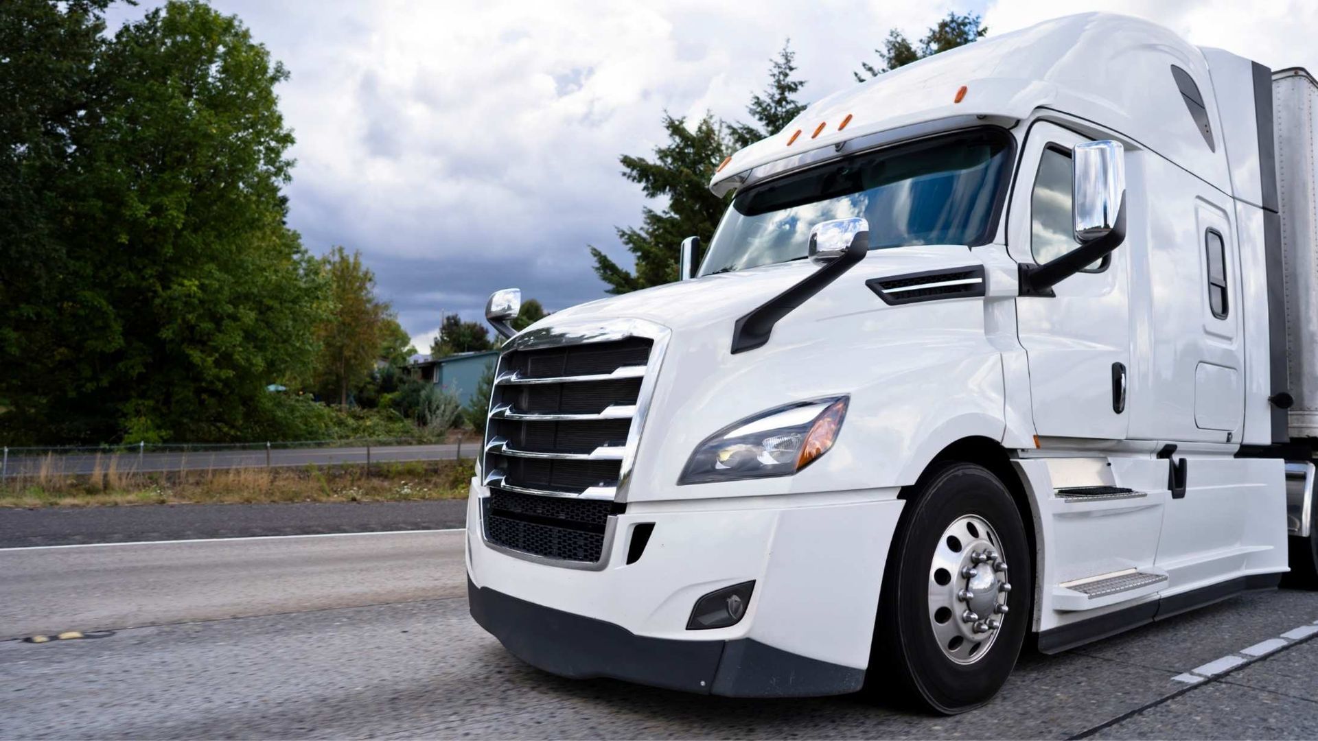 White semi-truck driving on a highway, trees in the background, overcast sky.