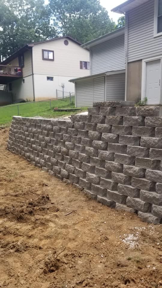 A retaining wall constructed of gray blocks, in front of two-story houses and a yard. The ground is dirt.