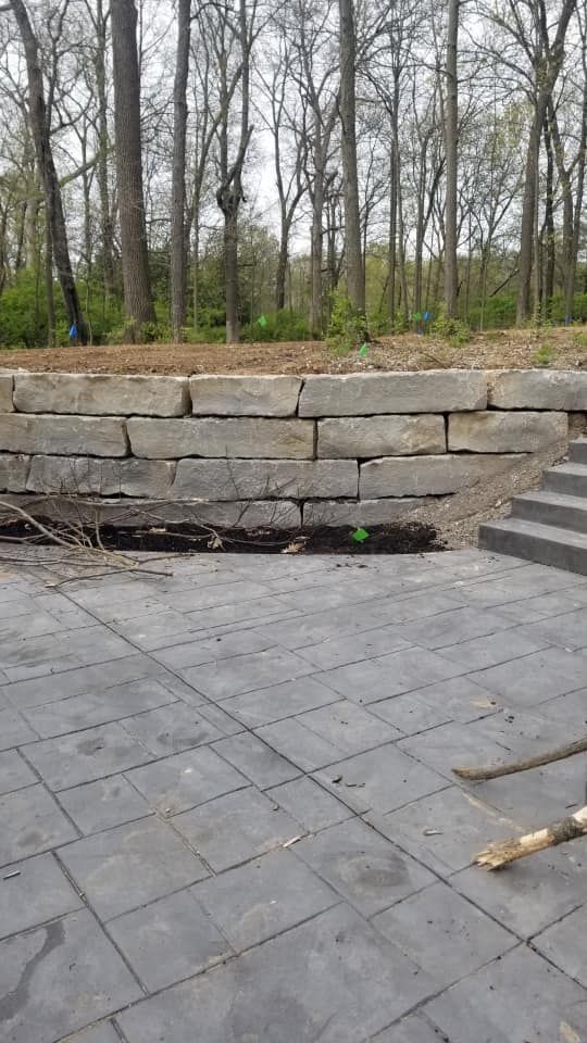 Stone retaining wall with a paved patio in the foreground, leading up to a set of stairs. Trees are in the background.
