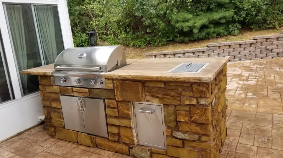 Outdoor kitchen island with a stainless steel grill, doors, and countertop; built of stone-like blocks, set on a patio.