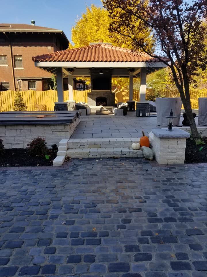 A stone patio with a gazebo featuring a fireplace, pumpkins, and a backdrop of autumn foliage and a house.