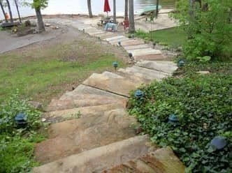 Stone steps lead down to a lakeside beach. Green grass and shrubs flank the steps, with pathway lights along the sides.