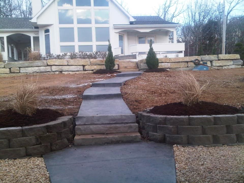 A paved walkway with steps leading to a white house. Dark stone walls border the walkway and landscaped flower beds.