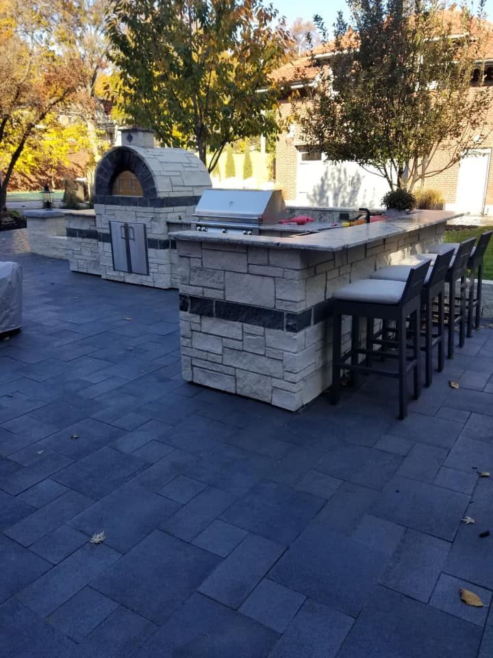 Outdoor kitchen with stone-clad counters, a pizza oven, grill, and bar seating. The patio has blue-gray paving.