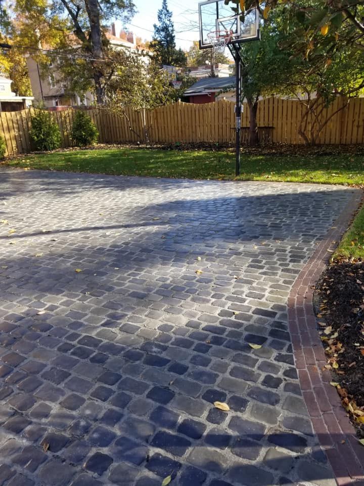 A black cobblestone driveway leading to a green lawn with a basketball hoop, bordered by a brick edge and a wooden fence.