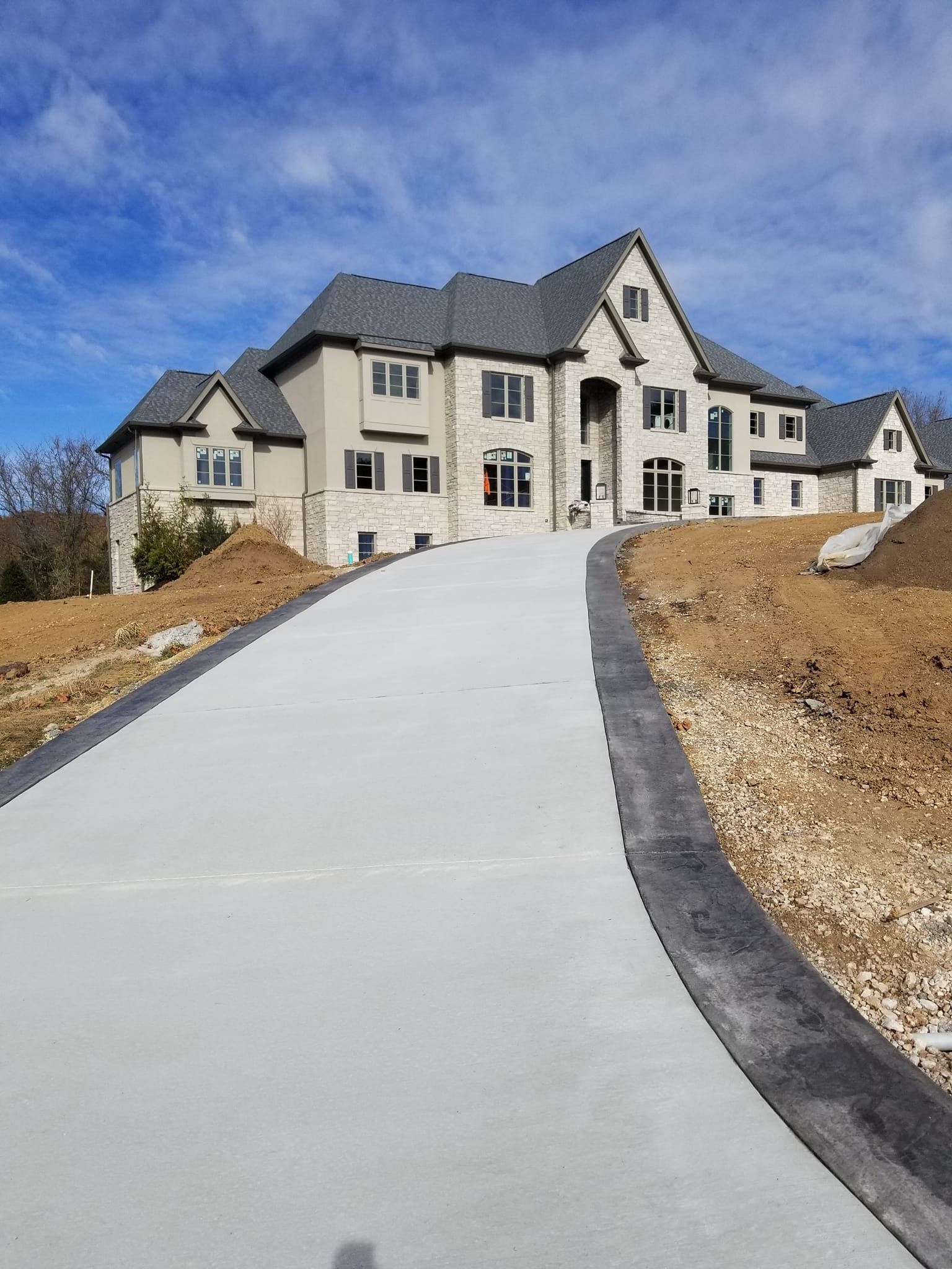 Long concrete driveway leading to a large two-story house with stone and stucco facade under a blue sky.