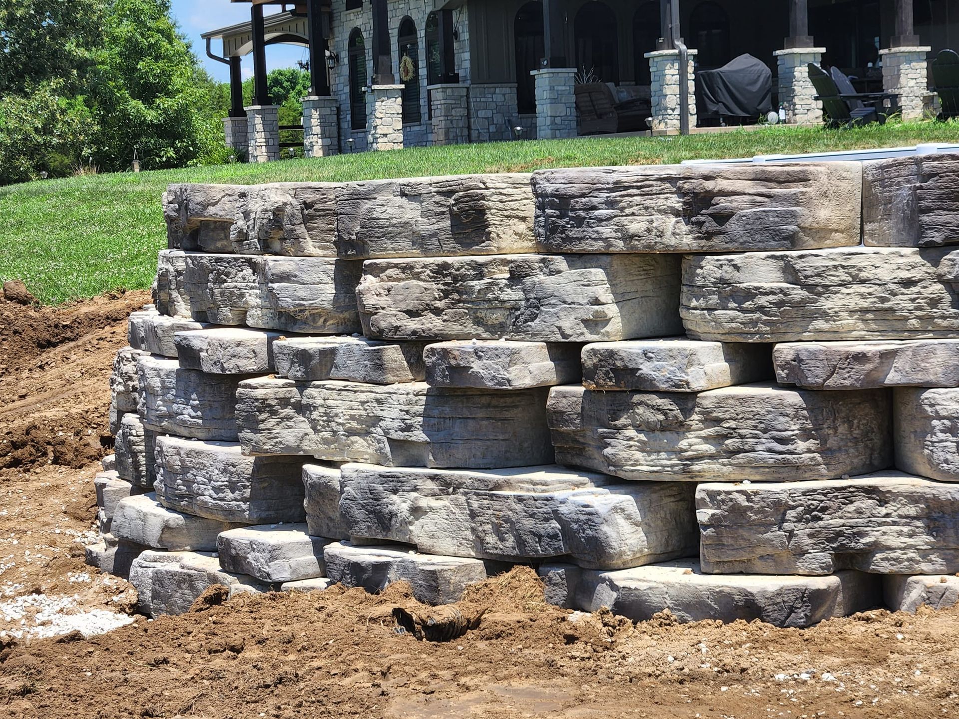 Stone retaining wall made of gray blocks, in front of a house with a grassy hill and surrounding dirt.