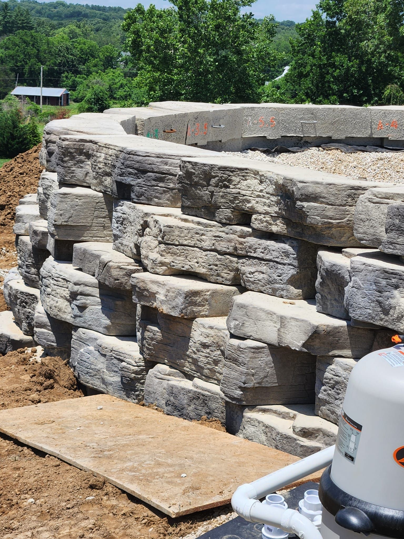 Stone retaining wall under construction, with blocks stacked in a curve. The setting is outdoors, near a pool pump.