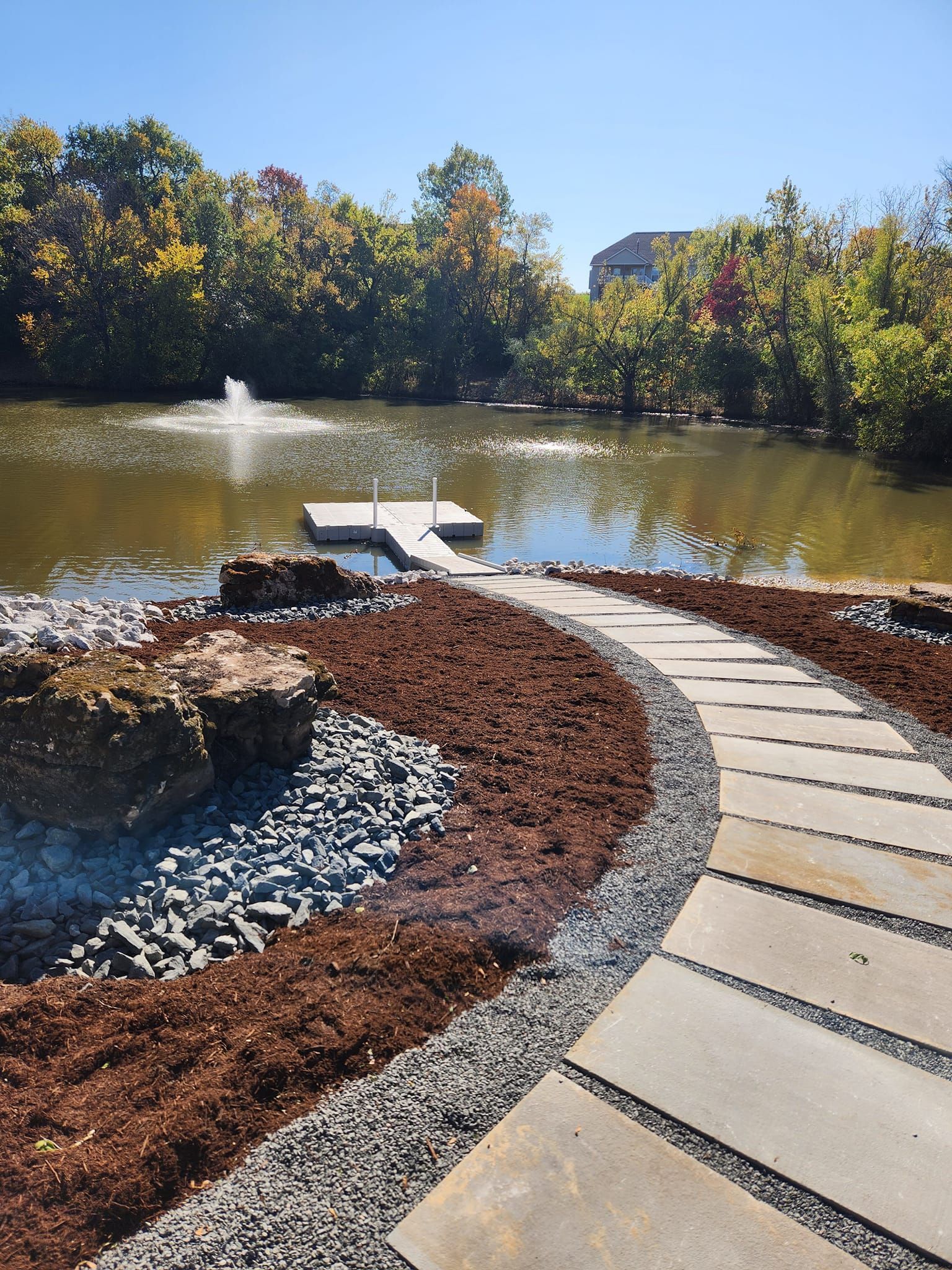 Curving stone path leads toward a small lake with a dock. Brown mulch, rocks, and trees surround the water under a blue sky.